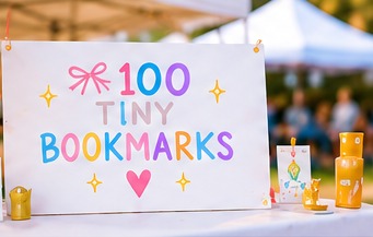 A 100 Tiny Bookmarks sign on a booth table at an outdoor community fair